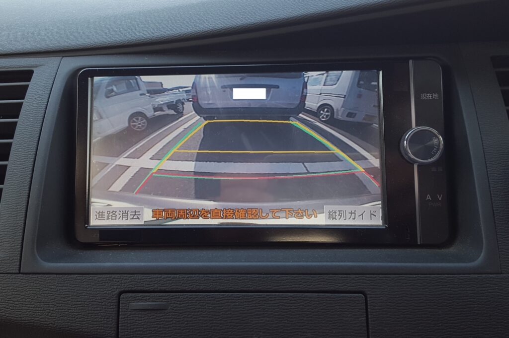 車のバックモニター画面。駐車スペースと後方の車両が映し出され、赤・黄・緑のガイドラインで距離感を示している。